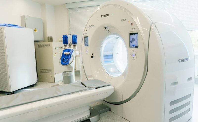 Moderns baltas krāsas Canon Aquilion ONE datortomogrāfijas aparāts ar pacienta gultu gaišā medicīnas kabinetā.