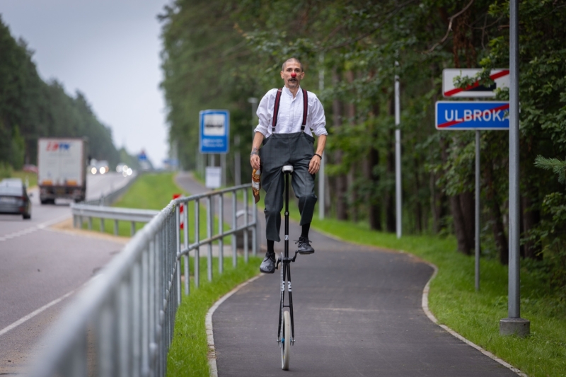 Persona brauc ar augstu vienriteni pa jaunizbūvētu gājēju un velobraucēju ceļu gar meža ieskautu autoceļu. Persona tērpusies skatuves mākslinieka stilam raksturīgā apģērbā un ar sarkanu degunu, demonstrējot līdzsvaru uz velosipēda. Fonā redzams autoceļš, ceļa zīmes, aizsargbarjeras un priežu mežs. Attēls uzņemts infrastruktūras objekta atklāšanas vai publiska pasākuma laikā.