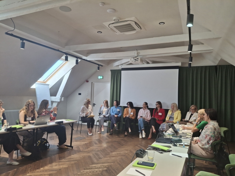 Semināra dalībnieki sēž aplī konferenču telpā ar slīpiem griestiem un projektora ekrānu priekšā.