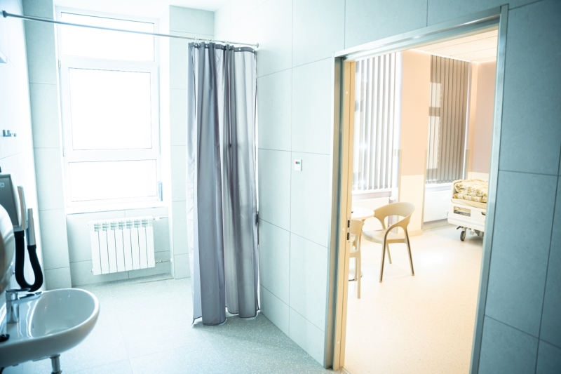 Mūsdienīga slimnīcas sanitārā telpa ar izlietni, dušu ar aizkaru un atvērtām durvīm uz palātu.