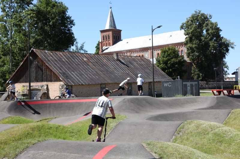 Bērni un jaunieši izmanto asfaltētu velotrasi jeb “pump track” trasi brīvā dabā. Priekšplānā redzams bērns ar skrejriteni, bet fonā vairāki bērni pārvietojas ar velosipēdiem un skrejriteņiem. Aiz trases atrodas vēsturiska mūra ēka un baznīca ar torni, apkārt aug koki. Attēls uzņemts saulainā vasaras dienā.
