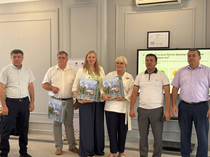 Oficiāla tikšanās telpās, kur seši dalībnieki pozē kopīgai fotogrāfijai. Divi centra dalībnieki rokās tur Uzbekistānas suvenīru dāvanu komplektus. Fonā redzams prezentācijas ekrāns ar uzbeku valodas tekstu un informatīvs stends.