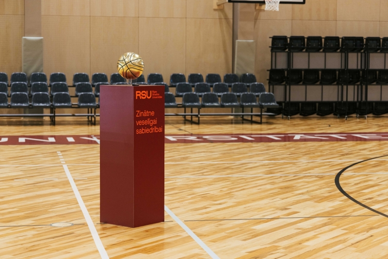 Jaunās sporta halles centrā uz pjedestāla novietota dekoratīva basketbola bumba. Apkārt redzams koka sporta laukuma segums, skatītāju tribīnes un basketbola grozs, kas raksturo modernas sporta infrastruktūras vidi.