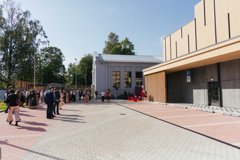 Pie Rīgas Stradiņa universitātes jaunā sporta kompleksa pulcējušies pasākuma apmeklētāji. Priekšplānā redzams plašs bruģēts laukums, bet labajā pusē moderna sporta ēkas fasāde. Fonā atrodas vēsturiska ēka un koki saulainā dienā.
