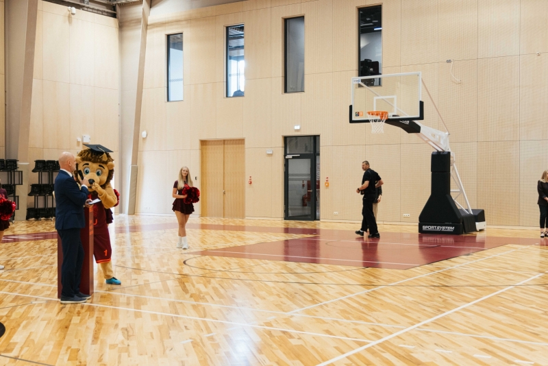 Sporta hallē notiek atklāšanas pasākums. Basketbola laukumā redzami pasākuma dalībnieki, talismans un karsējmeitenes. Plašā zāle aprīkota ar basketbola grozu, tribīnēm un mūsdienīgu sporta infrastruktūru.