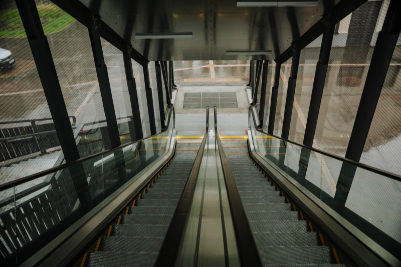 Skats no stiklotas gājēju pārejas uz leju pa eskalatoru. Modernā infrastruktūra nodrošina ērtu un pieejamu nokļūšanu starp dažādiem transporta veidiem un pilsētvides objektiem.