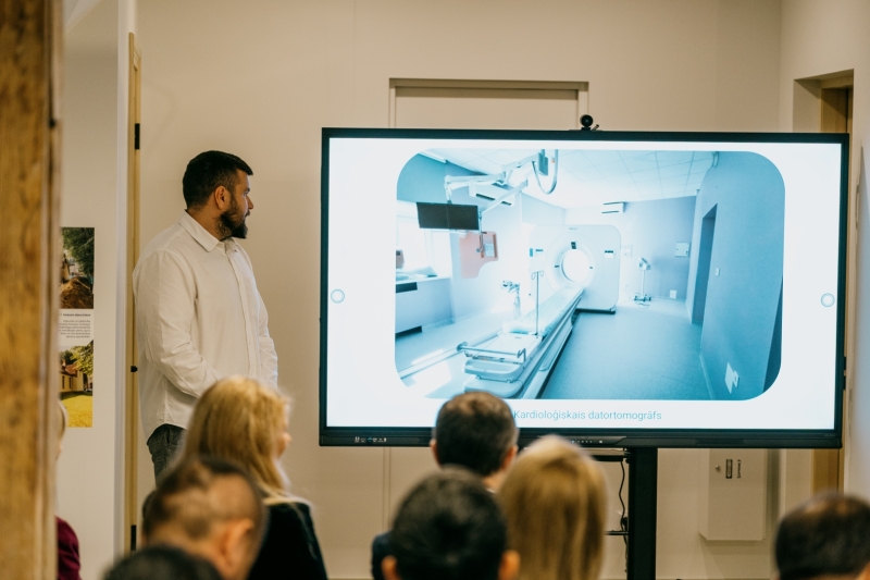Dalībnieki klausās prezentāciju medicīnas iestādes telpā. Pie liela ekrāna stāv prezentētājs, bet uz ekrāna redzams datortomogrāfijas izmeklējumu kabinets ar medicīnisko aprīkojumu un datortomogrāfu.