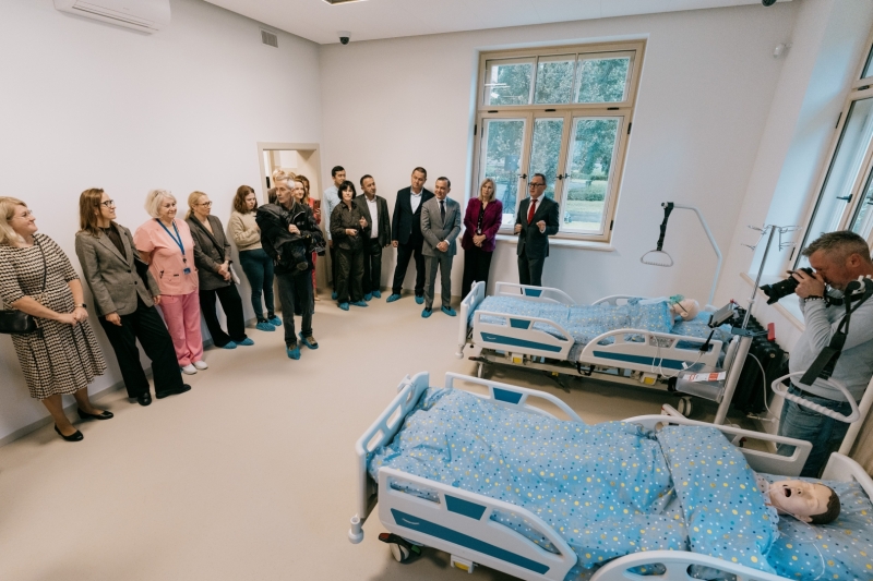 Slimnīcas palātā grupa cilvēku aplūko moderni aprīkotu pacientu aprūpes telpu. Telpā atrodas vairākas slimnīcas gultas ar manekeniem, medicīniskais aprīkojums un fotogrāfs, kurš dokumentē notikumu.