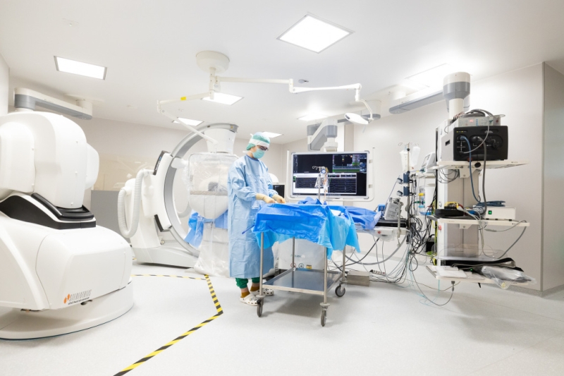 Medicīnas speciālists procedūru telpā pie galda, ko ieskauj robotizēta medicīnas iekārta, monitori un cita ārstniecības aparatūra.