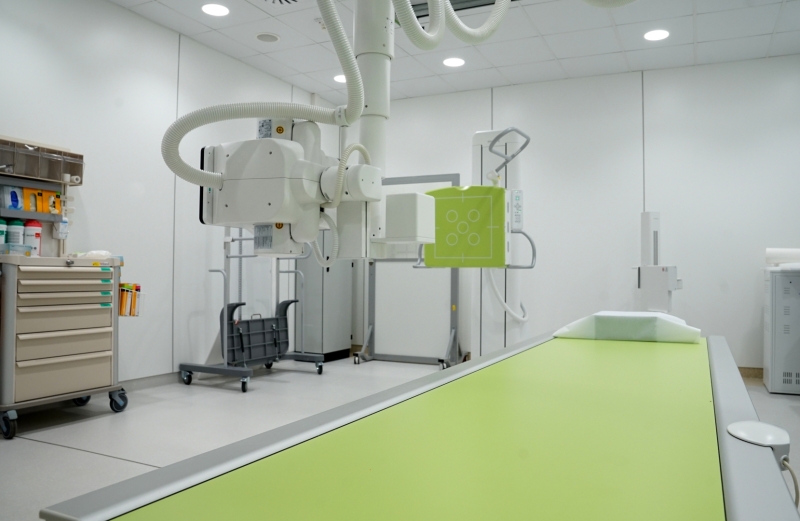 Rentgena izmeklējumu telpa ar diagnostikas iekārtu un zaļas krāsas pacienta galdu.