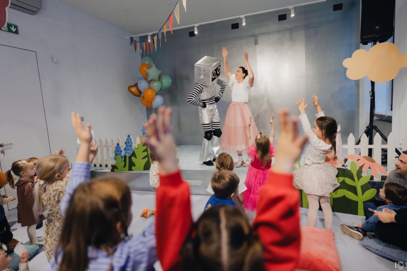 Bērni bērnu pasākumā paceļ rokas un iesaistās rotaļā kopā ar animatoriem robota un princeses kostīmos.