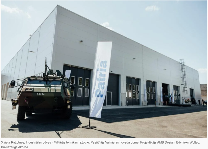 Liela, moderna rūpnieciska ēka ar vairākām garāžas durvīm, priekšplānā militārs transportlīdzeklis ar identifikatoru "AA514". Blakus transportlīdzeklim novietots balts karogs ar uzrakstu "Patria" zilā krāsā. Attēls uzņemts dienas laikā zem skaidrām debesīm, norādot uz aizsardzības vai ražošanas aktivitātēm, kas saistītas ar Patria uzņēmumu.
