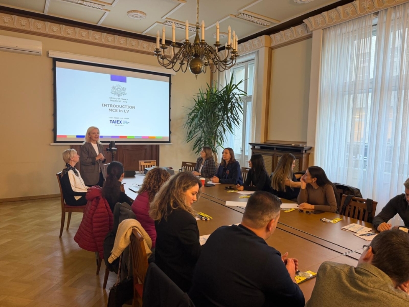 Starptautiskas pieredzes apmaiņas vizītes dalībnieki sēž pie konferenču galda vēsturiskā sanāksmju telpā un klausās prezentāciju. 