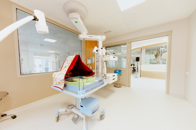 Gaiša un moderna slimnīcas telpa ar jaundzimušo aprūpes gultiņu un medicīnisko aprīkojumu.
