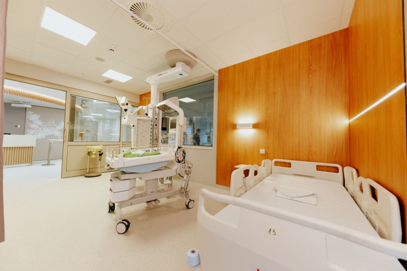 Slimnīcas palāta ar pacienta gultu, jaundzimušo aprūpes gultiņu un medicīnisko aprīkojumu.