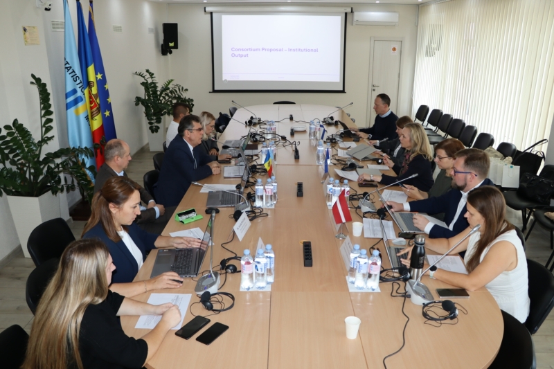Cilvēku grupa sēž ap garu sanāksmju galdu konferenču telpā darba sanāksmes laikā, uz galda izvietoti klēpjdatori, mikrofoni un Moldovas un Latvijas karodziņi.