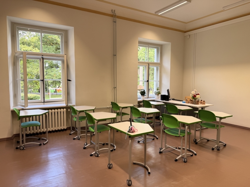 Atjaunota mācību klase ar gaišām sienām, lieliem logiem un zaļiem pārvietojamiem galdiem un krēsliem. Klases priekšpusē atrodas skolotāja darba vieta ar datoru, uz palodzēm novietoti telpaugi. Pa logiem redzami koki un apstādījumi.