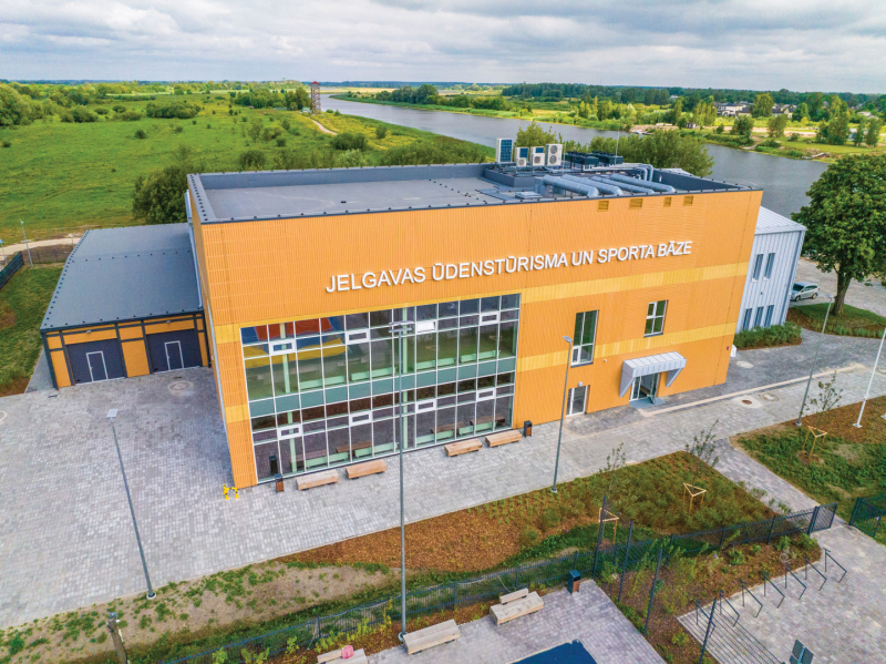 Aerofotoattēls ar mūsdienīgu ēku, uz kuras fasādes redzams uzraksts "JELGAVAS ŪDENSTŪRISMA UN SPORTA BĀZE". Ēkai ir dzeltenpelēka apdare un lieli logi. Tā atrodas zaļā, atklātā teritorijā pie ūdenskrātuves, apkārt redzami zālāji, koki un celiņi, kas ved uz ūdeni. Attēls ataino vietu, kas paredzēta aktīvai atpūtai un sportam pie ūdens.
