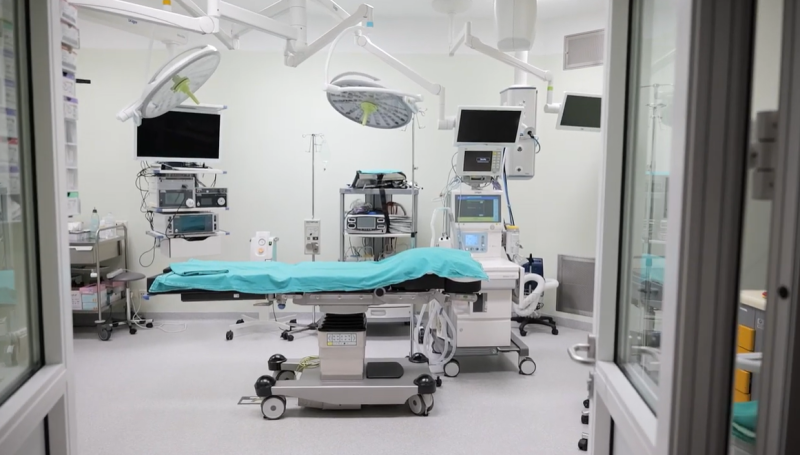 Mūsdienīga slimnīcas operāciju zāle ar operāciju galdu telpas centrā. Virs galda izvietotas vairākas operāciju lampas, bet apkārt redzamas medicīniskās iekārtas, monitori un anestēzijas aprīkojums. Telpa ir gaiša, tīra un sagatavota pacientu uzņemšanai.