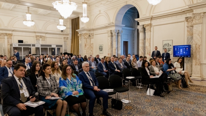 Konferences zāle ar augstiem griestiem un lustru apgaismojumu, kur rindās sēž pasākuma dalībnieki, klausoties prezentāciju; zāles labajā pusē redzams ekrāns ar vizuālo materiālu.