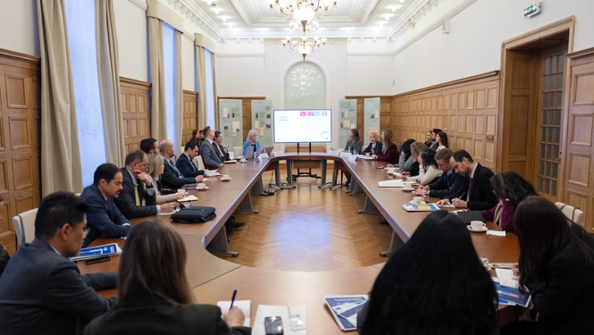 Sanāksmju telpā pie U veida galda notiek plaša darba tikšanās ar prezentāciju ekrānā priekšplānā.