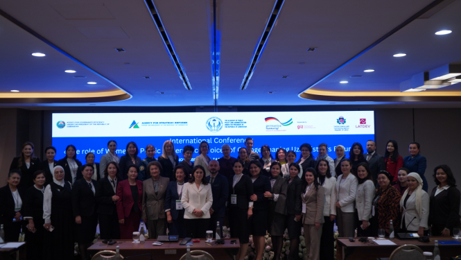 Grupas foto no starptautiskās konferences par dzimumlīdztiesību valsts pārvaldē Taškentā; dalībnieki pozē kopā konferenču zālē, fonā redzams liels ekrāns ar konferences nosaukumu un partneru logotipiem.