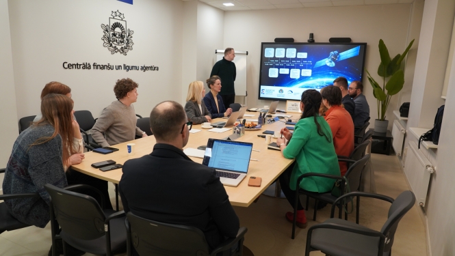 CFLA telpās notiek darba sanāksme – pie galda sēž projekta partneri, priekšā redzams ekrāns ar prezentāciju par satelītu risinājumiem un digitālo savienojamību