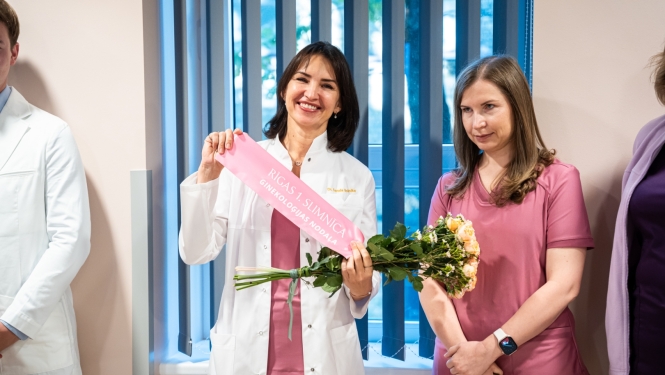 Divas mediķes pasākumā: ārste baltā uzsvārcī tur ziedu pušķi ar rozā lentu "Rīgas 1. slimnīca Ginekoloģijas nodaļa", blakus stāv kolēģe sārtā medicīniskajā tērpā.