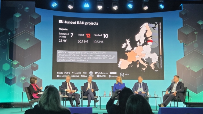 Paneļdiskusija tehnoloģiju konferencē. Uz skatuves pieci diskusijas dalībnieki un moderators, bet lielajā ekrānā redzama prezentācija par ES finansētiem pētniecības un attīstības projektiem Eiropā.