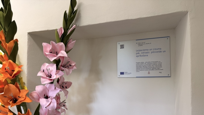 Pie sienas nišā izvietota informatīva plāksne par Eiropas Savienības līdzfinansētu projektu “Lejasciema un Lizuma psk. infrastr. pilnveide un aprīkošana”. Blakus plāksnei novietoti dekoratīvi rozā un oranži ziedi, telpā redzams radiators un gaišas krāsas siena.
