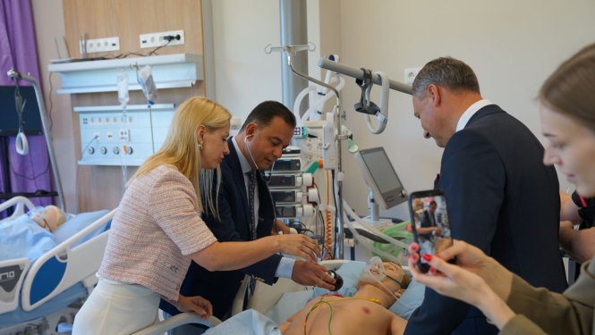 Vairākas personas simulāciju centra palātā iepazīstas ar medicīnisko mācību manekenu, kas novietots slimnīcas gultā. Telpā redzamas pacientu aprūpei paredzētas iekārtas, monitora ekrāni un cits medicīniskais aprīkojums. Viena persona notiekošo fotografē ar viedtālruni.