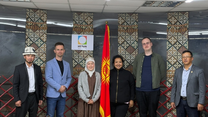 Sešu cilvēku grupa stāv rindā telpā pie Kirgizstānas karoga un sienas ar tradicionālajiem ziemeļnieku/kirgīzu rakstu panno.