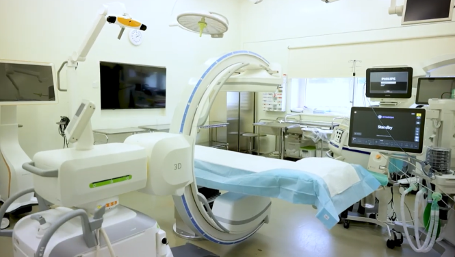 Slimnīcas operāciju zāle ar angiogrāfijas jeb rentgena izmeklējumu iekārtu, kuras lokveida konstrukcija atrodas virs procedūru galda. Apkārt izvietoti monitori, medicīniskās ierīces un aprīkojums pacientu uzraudzībai un ārstēšanai. Telpa ir labi apgaismota un aprīkota ar modernām tehnoloģijām.
