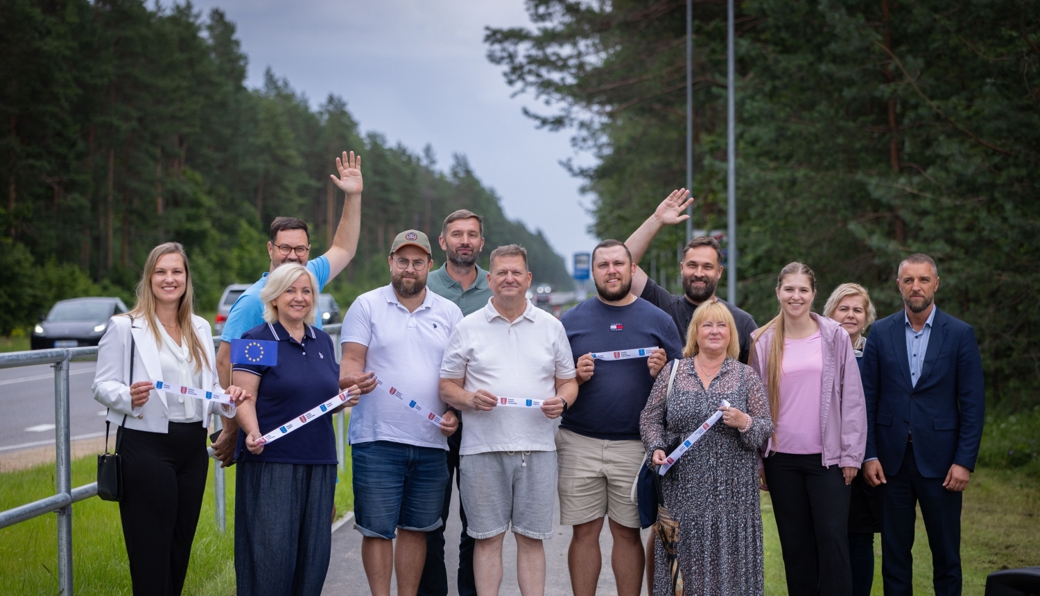 Grupa cilvēku stāv uz jauna gājēju un velosipēdu celiņa meža ieskautā ceļa malā. Vairāki dalībnieki rokās tur simbolisku atklāšanas lentu ar Eiropas Savienības un pašvaldības simboliku. Fonā redzama brauktuve, koki un ceļa apgaismojums. Foto uzņemts infrastruktūras objekta atklāšanas pasākuma laikā brīvā dabā.