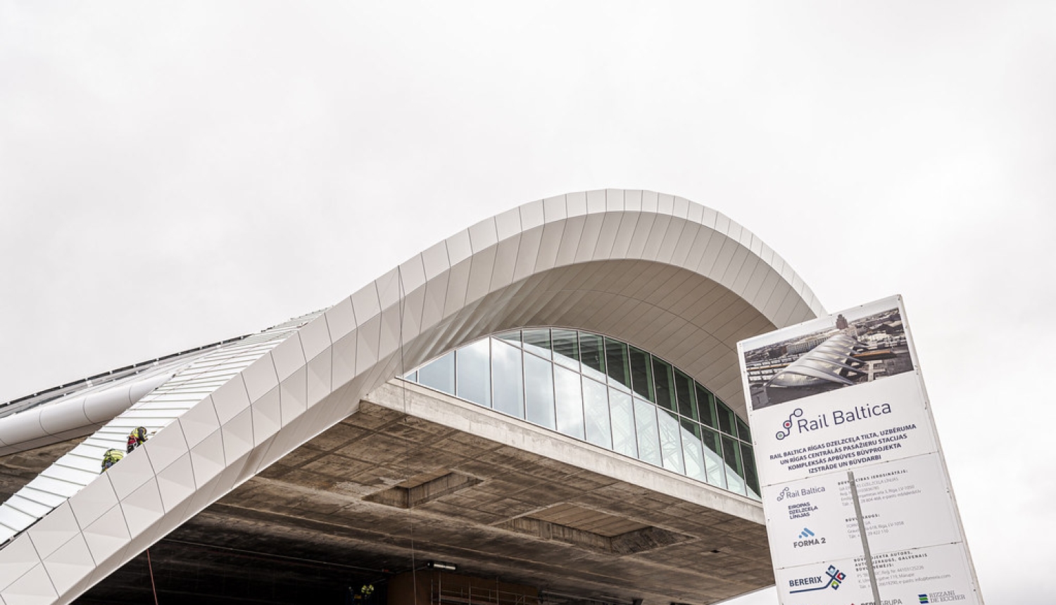 Rail Baltica Rīgas Centrālās stacijas ēkas būvniecība. Attēlā redzama stacijas iespaidīgā viļņveida jumta konstrukcija ar stiklotu fasādi. Priekšplānā izvietots projekta informatīvais stends, bet uz konstrukcijas un zem tās redzami būvdarbu veicēji.