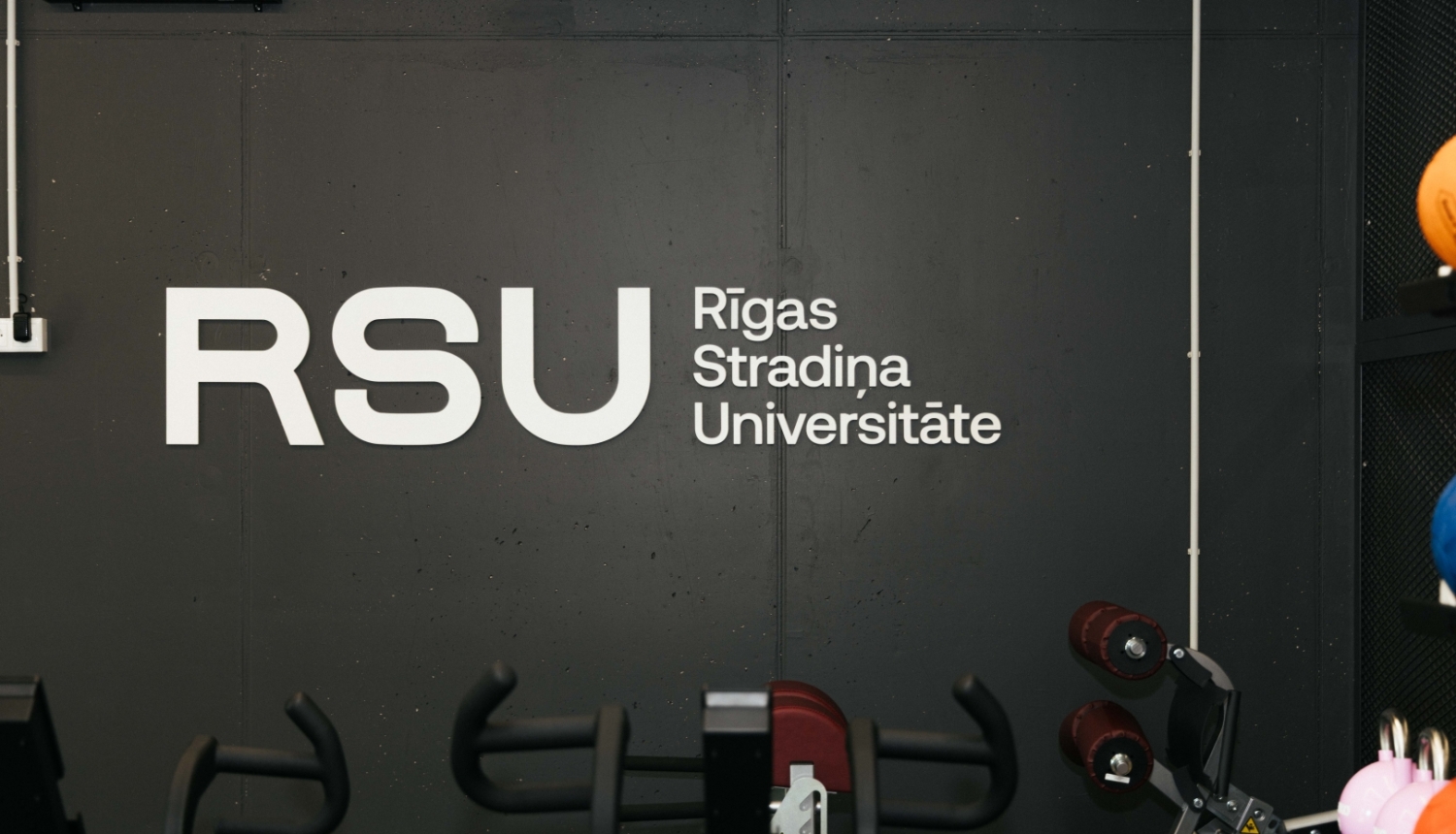 Trenažieru zālē redzams Rīgas Stradiņa universitātes logotips uz tumšas sienas. Priekšplānā izvietoti dažādi fitnesa trenažieri un sporta inventārs, kas paredzēts studentu, sportistu un pētnieku vajadzībām.