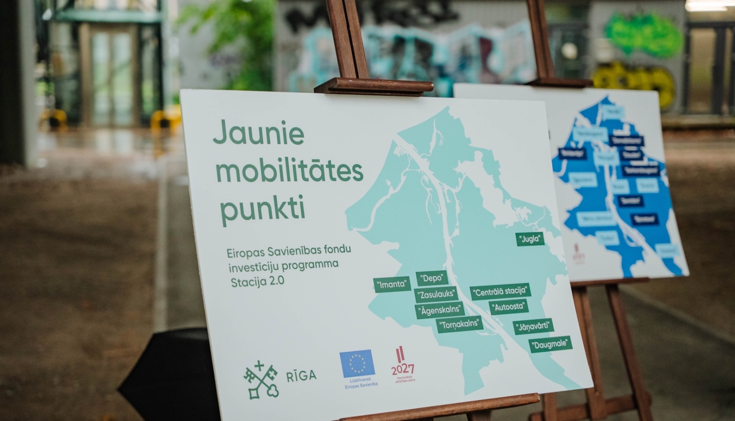 Informatīvs stends uz molberta ar uzrakstu “Jaunie mobilitātes punkti” un Rīgas karti, kurā atzīmētas dzelzceļa staciju apkārtnēs izbūvētās mobilitātes punktu atrašanās vietas.
