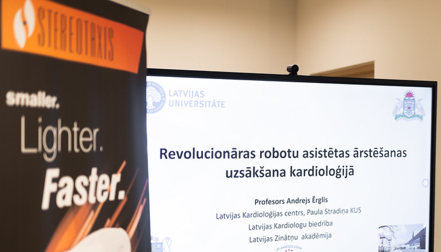 Ekrānā redzama prezentācija “Revolucionāras robotu asistētas ārstēšanas uzsākšana kardioloģijā”; priekšplānā daļēji redzams Stereotaxis stends.