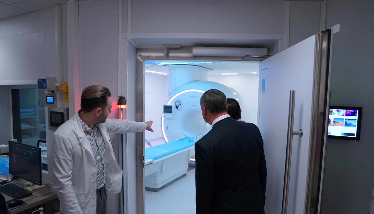Medicīnas speciālists baltā virsvalkā demonstrē apmeklētājiem magnētiskās rezonanses iekārtu izmeklējumu telpā.