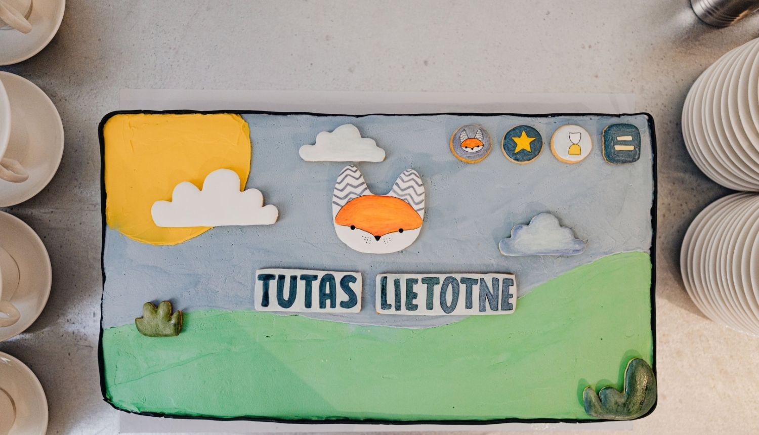 Bērnu pasākuma tematiskā kūka ar lapsas attēlu, sauli, mākoņiem un uzrakstu “Tutas lietotne”.