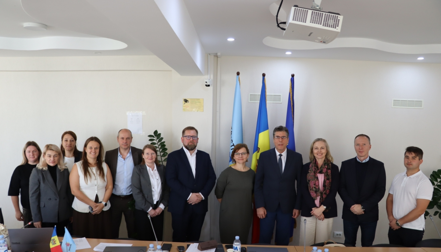Oficiāls delegācijas kopportrets — 11 cilvēki stāv rindā sanāksmju telpā iepretim valsts karogiem.
