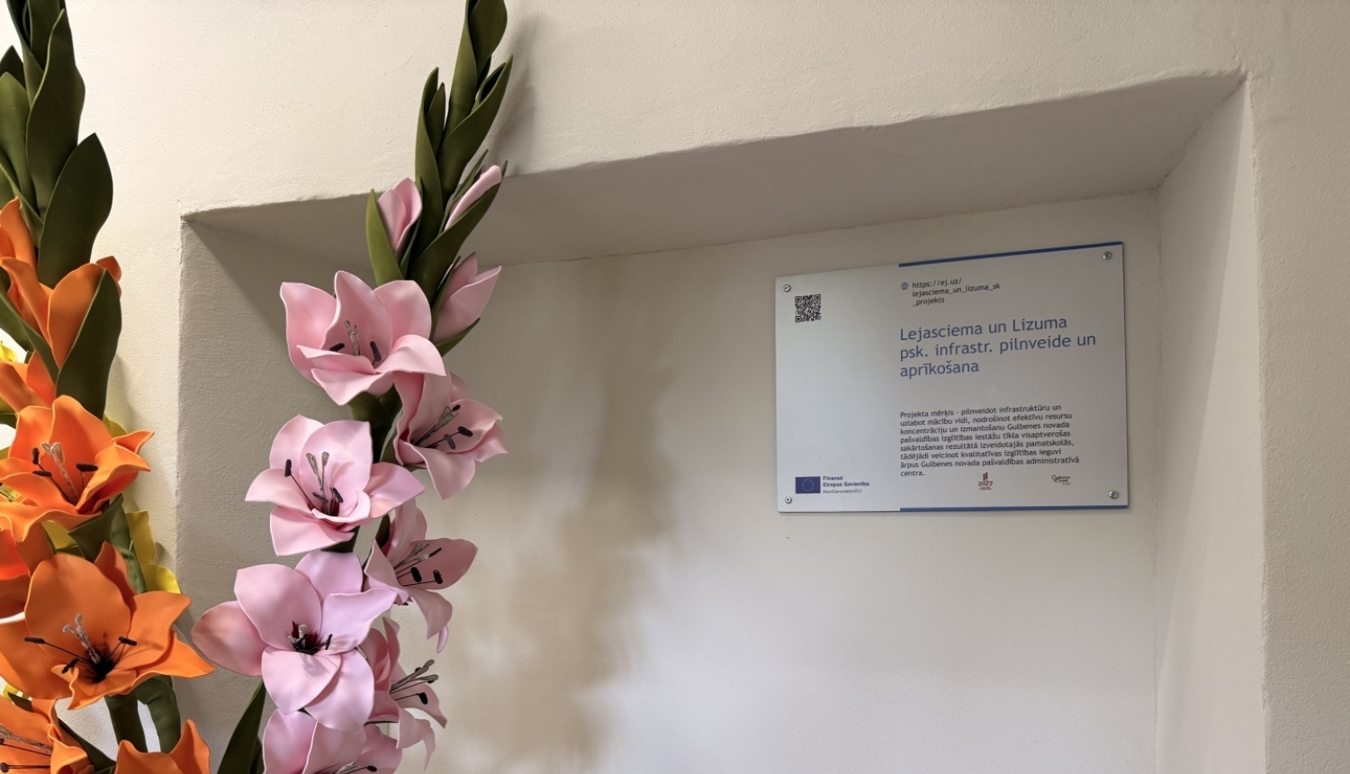 Pie sienas nišā izvietota informatīva plāksne par Eiropas Savienības līdzfinansētu projektu “Lejasciema un Lizuma psk. infrastr. pilnveide un aprīkošana”. Blakus plāksnei novietoti dekoratīvi rozā un oranži ziedi, telpā redzams radiators un gaišas krāsas siena.