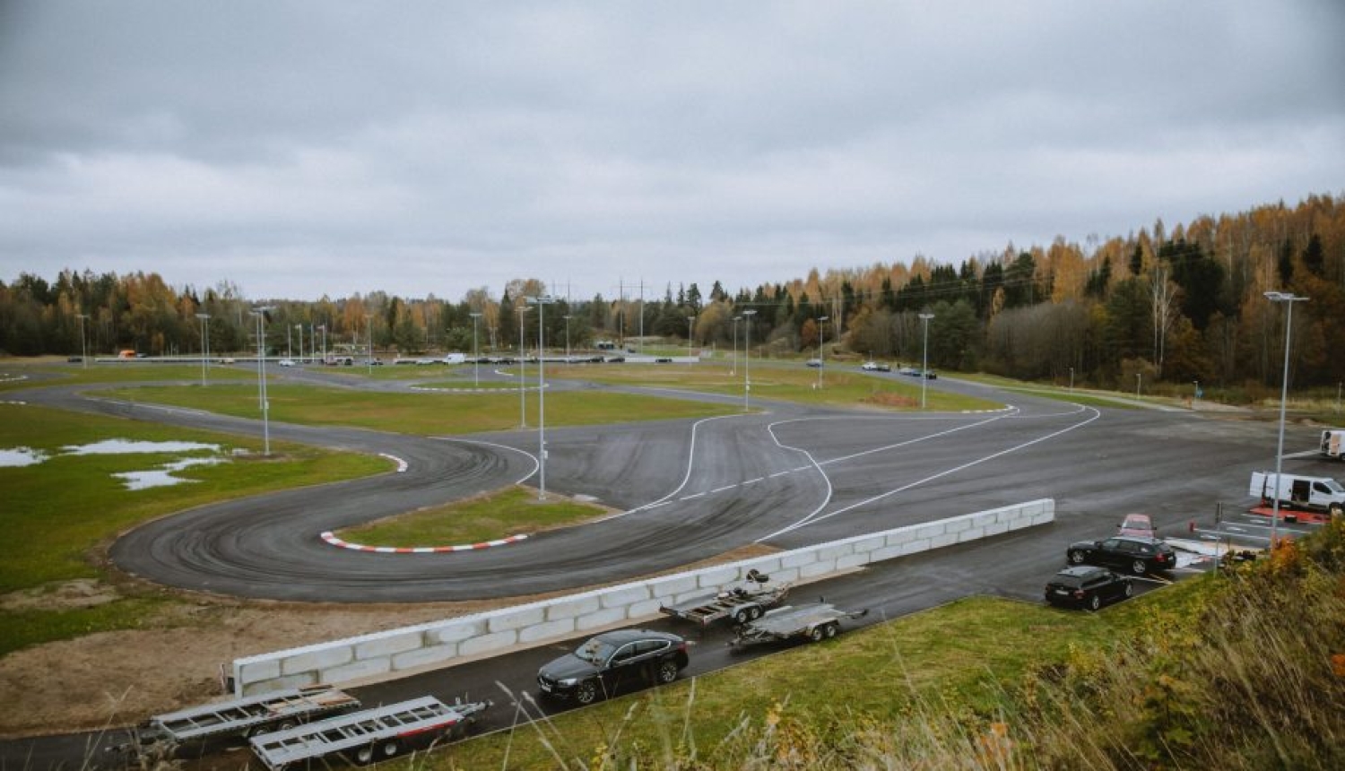 Plašs asfaltēts autosporta un braukšanas mācību laukums ar līkumiem un ceļa marķējumu, ko ieskauj rudens ainava.