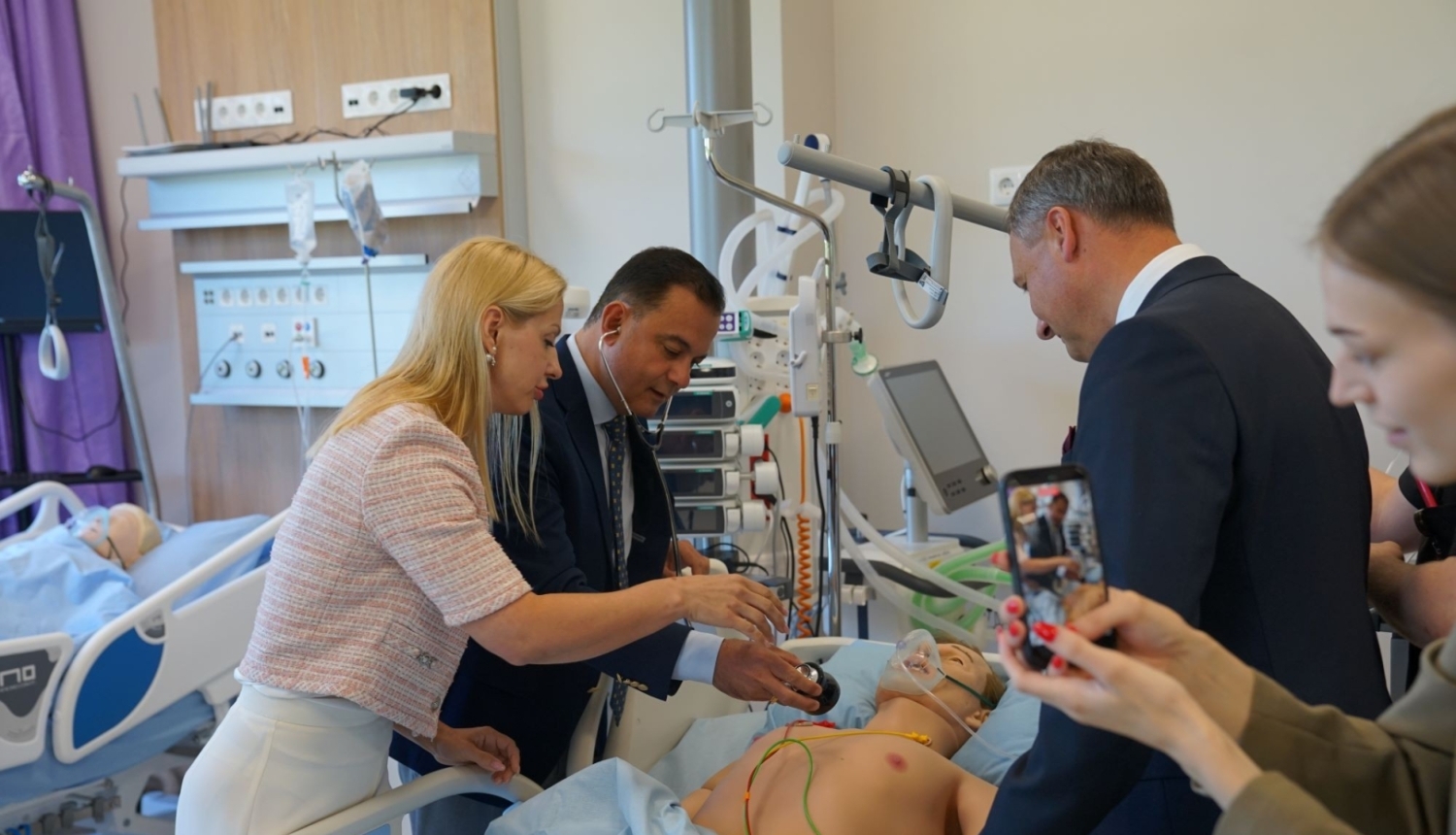Vairākas personas simulāciju centra palātā iepazīstas ar medicīnisko mācību manekenu, kas novietots slimnīcas gultā. Telpā redzamas pacientu aprūpei paredzētas iekārtas, monitora ekrāni un cits medicīniskais aprīkojums. Viena persona notiekošo fotografē ar viedtālruni.