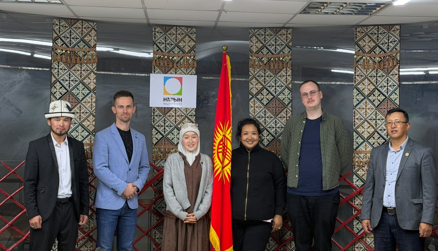 Sešu cilvēku grupa stāv rindā telpā pie Kirgizstānas karoga un sienas ar tradicionālajiem ziemeļnieku/kirgīzu rakstu panno.