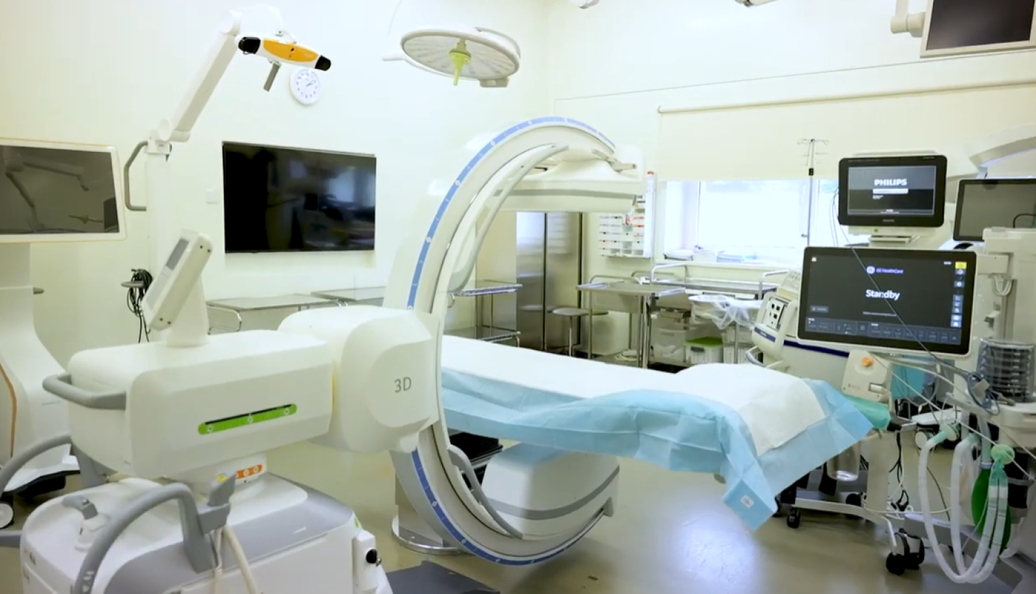 Slimnīcas operāciju zāle ar angiogrāfijas jeb rentgena izmeklējumu iekārtu, kuras lokveida konstrukcija atrodas virs procedūru galda. Apkārt izvietoti monitori, medicīniskās ierīces un aprīkojums pacientu uzraudzībai un ārstēšanai. Telpa ir labi apgaismota un aprīkota ar modernām tehnoloģijām.
