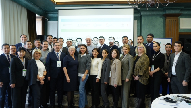 Liela profesionāļu grupa formālā apģērbā pozē kopbildē konferenču zālē; fonā redzams projektora ekrāns, lustras un pasākuma baneri.
