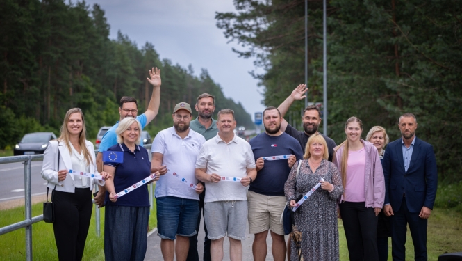 Grupa cilvēku stāv uz jauna gājēju un velosipēdu celiņa meža ieskautā ceļa malā. Vairāki dalībnieki rokās tur simbolisku atklāšanas lentu ar Eiropas Savienības un pašvaldības simboliku. Fonā redzama brauktuve, koki un ceļa apgaismojums. Foto uzņemts infrastruktūras objekta atklāšanas pasākuma laikā brīvā dabā.