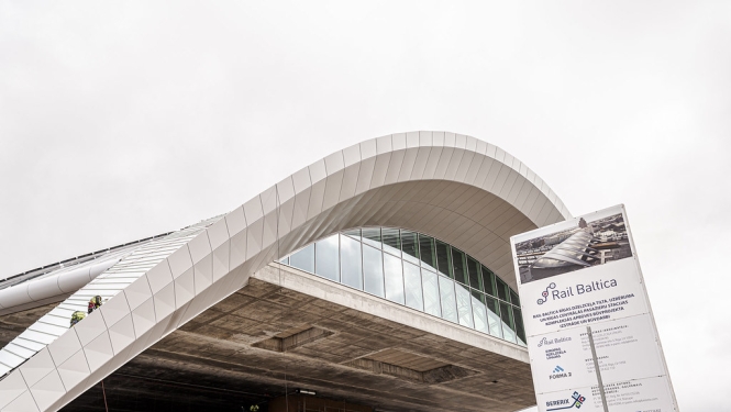 Rail Baltica Rīgas Centrālās stacijas ēkas būvniecība. Attēlā redzama stacijas iespaidīgā viļņveida jumta konstrukcija ar stiklotu fasādi. Priekšplānā izvietots projekta informatīvais stends, bet uz konstrukcijas un zem tās redzami būvdarbu veicēji.