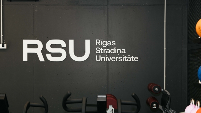 Trenažieru zālē redzams Rīgas Stradiņa universitātes logotips uz tumšas sienas. Priekšplānā izvietoti dažādi fitnesa trenažieri un sporta inventārs, kas paredzēts studentu, sportistu un pētnieku vajadzībām.